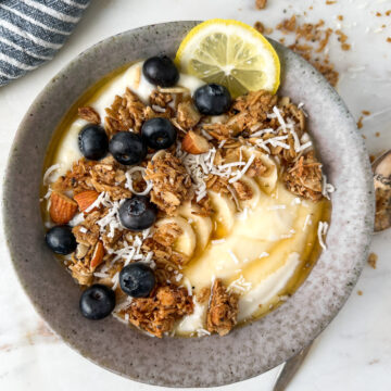 Granola and lemon yogurt bowl covered in blueberries with a spoon and granola crumbs on a table.