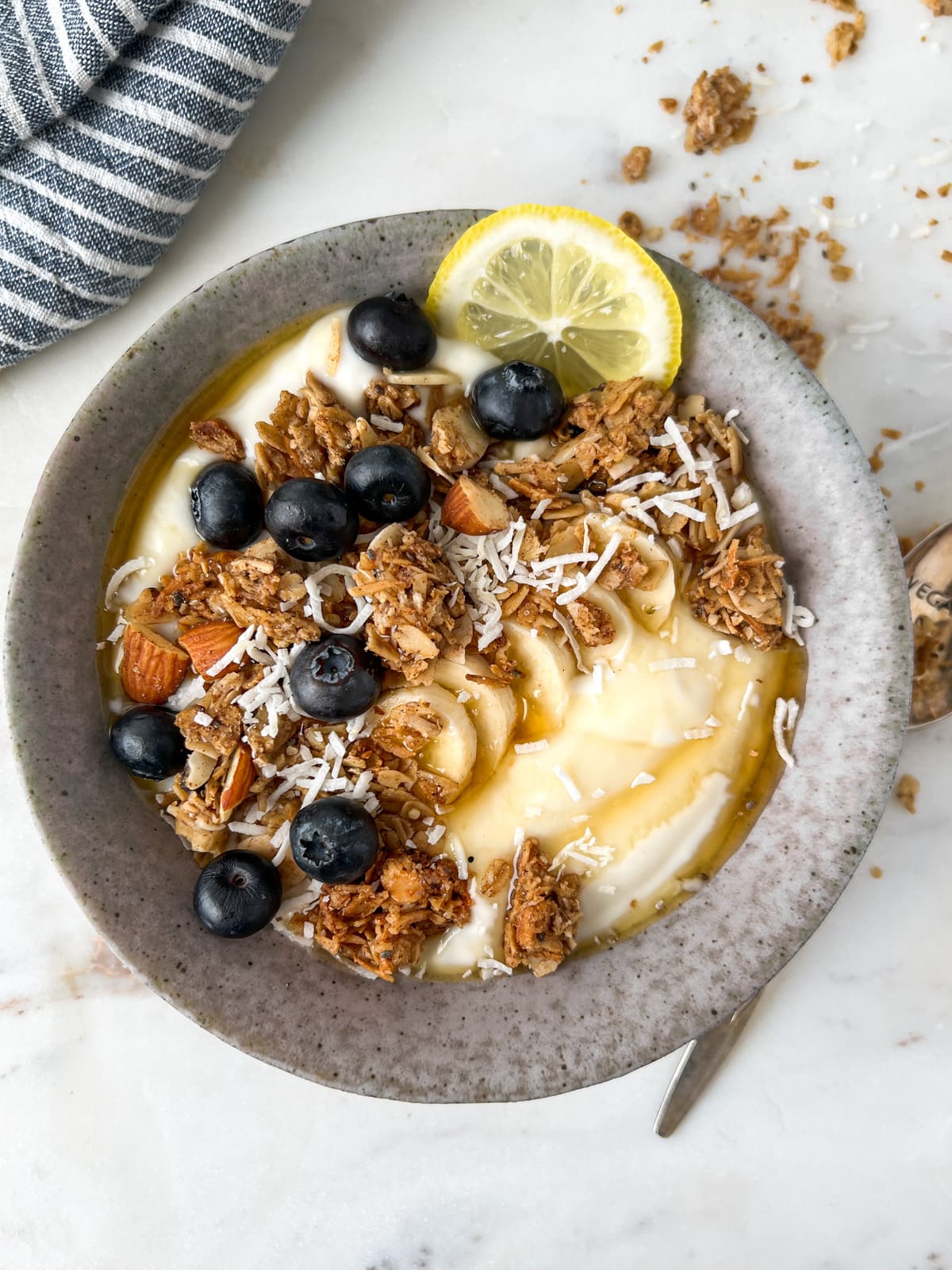 Granola and lemon yogurt bowl covered in blueberries with a spoon and granola crumbs on a table.