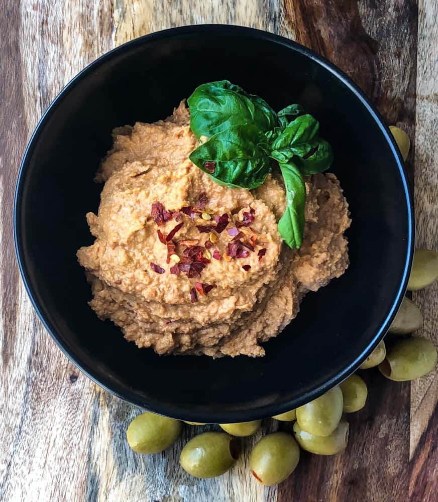 Tangy Tomato and Olive Hummus in a bowl surrounded by olives, garnished with a sprig of basil and sprinkle of hot pepper flakes