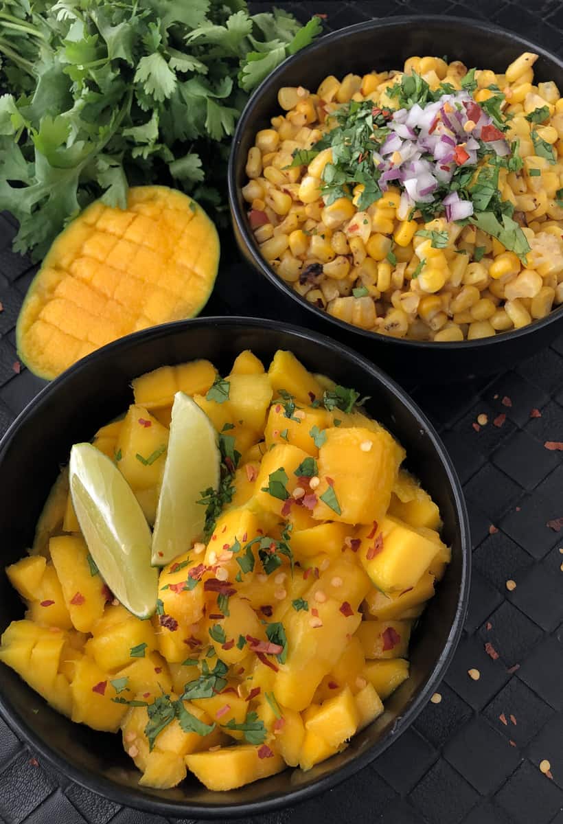 Bowls full of chopped mango and grilled corn on a cutting board.