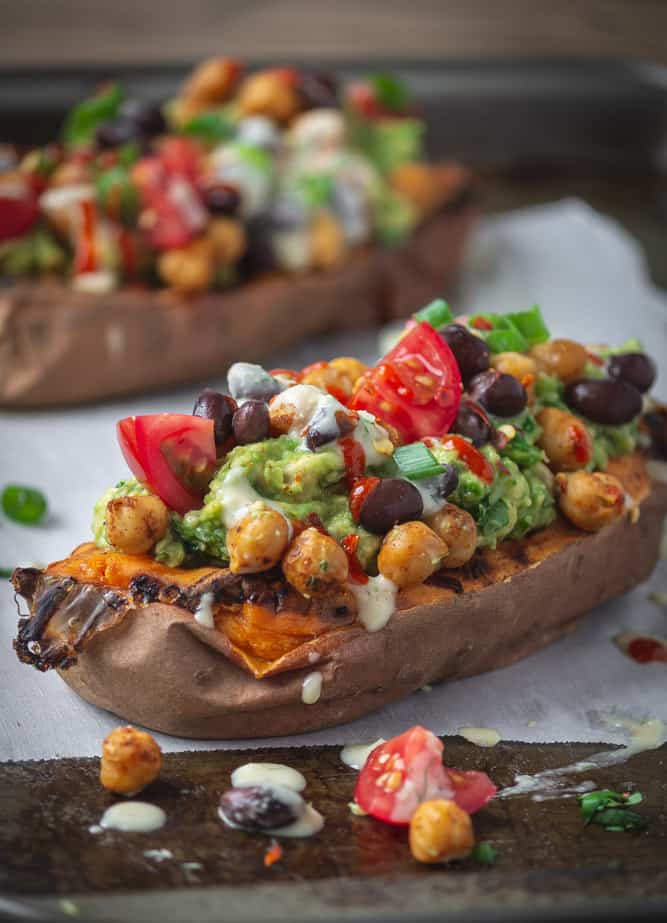 Close up of stuffed sweet potato with black beans, guacamole, tomatoes and drizzled with hummus and sriracha served on a baking sheet.