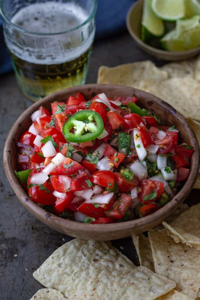 Bowlful of pico de gallo topped with a slice of jalapeno and surrounded by a pile of chips.