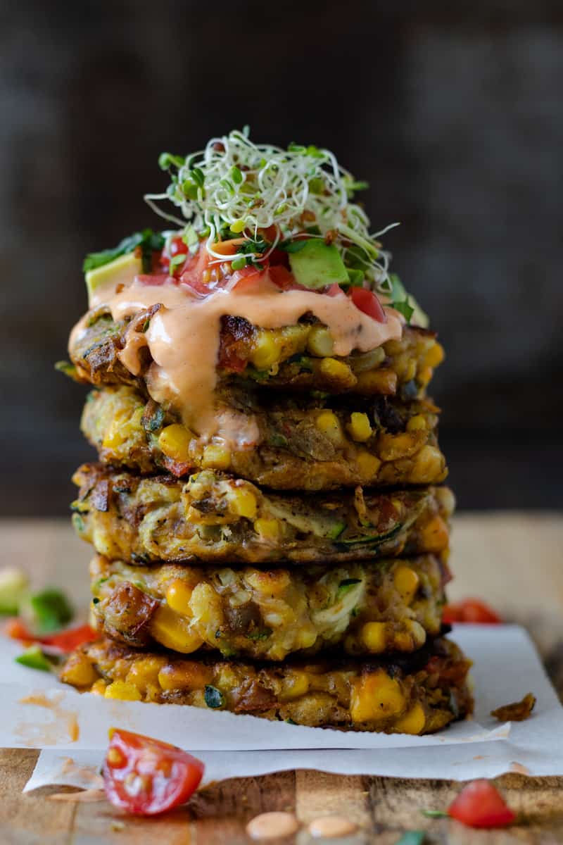 Stack of vegan corn fritters topped with tomatoes avocado sprouts and sauce.