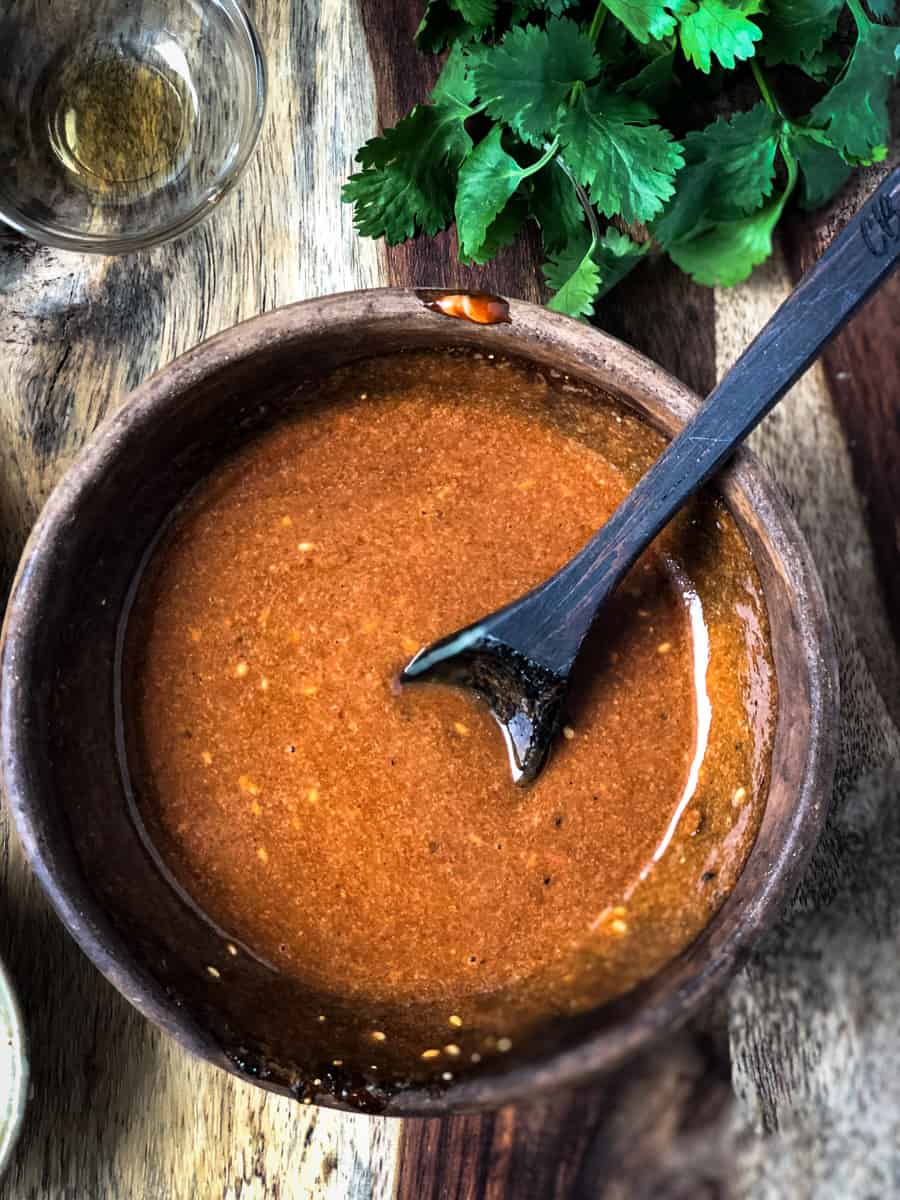 Spicy sauce in a bowl with a spoon and herbs scattered around the bowl.