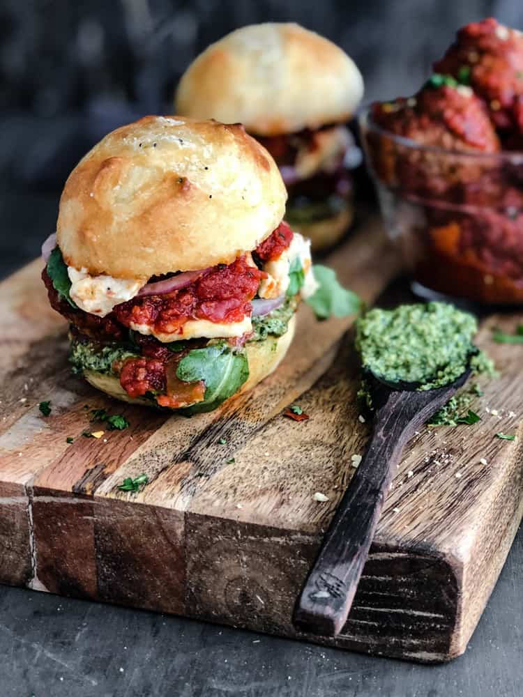 Crusty buns filled with meatballs, marinara sauce, cheese, and pesto on sitting on a cutting board.