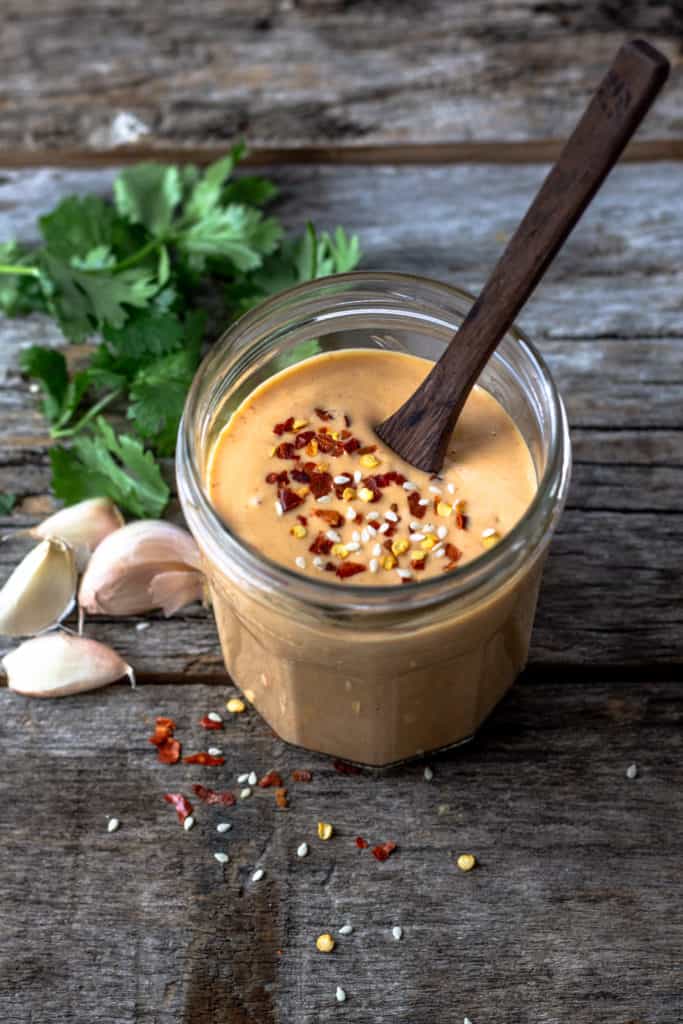 Thick orange spicy tahini sauce in a jar with a wooden spoon for serving and sprinkled with red pepper flakes.