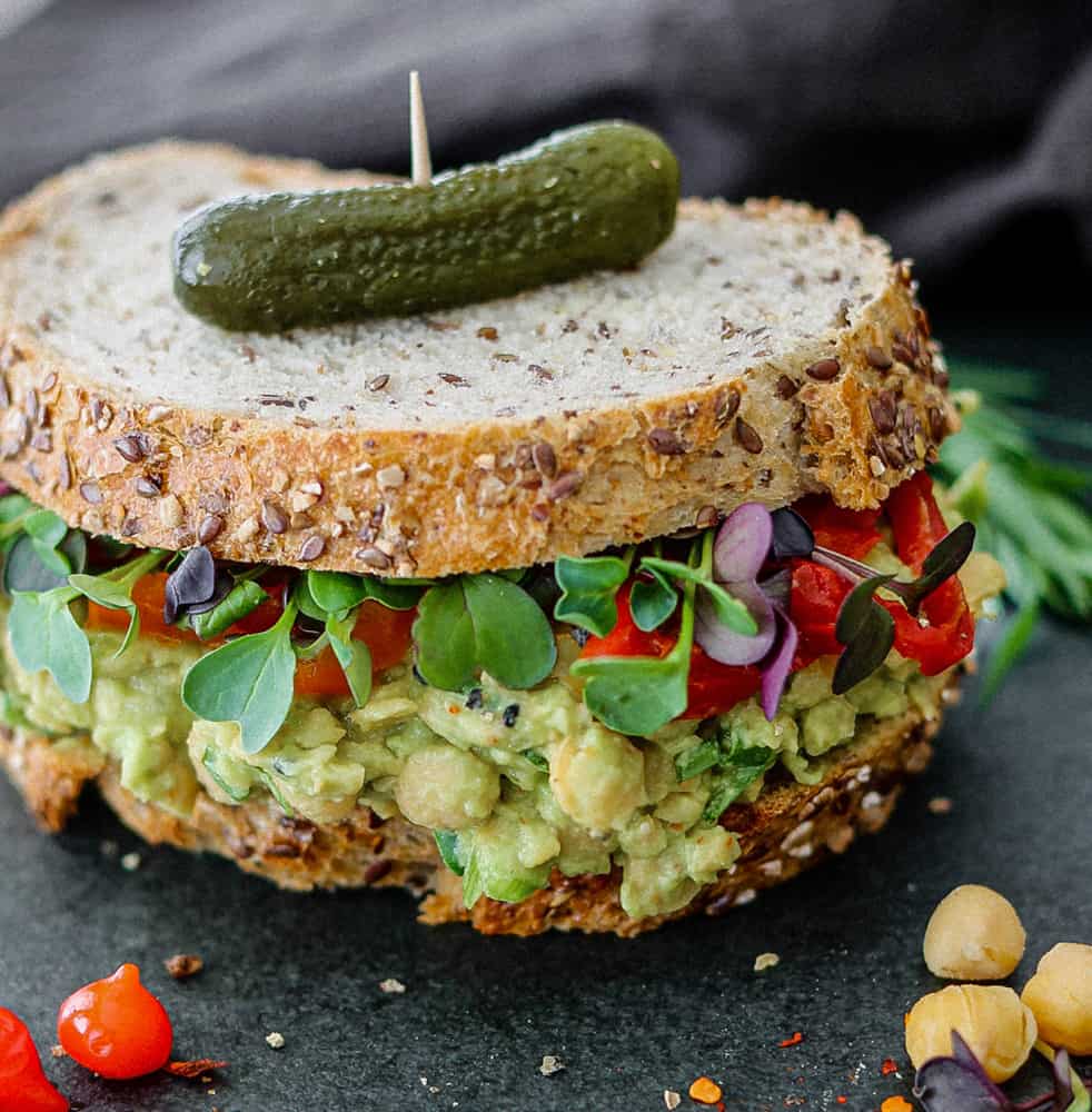 Chickpea avocado sandwich on grainy bread with sprouts and pickle on top.