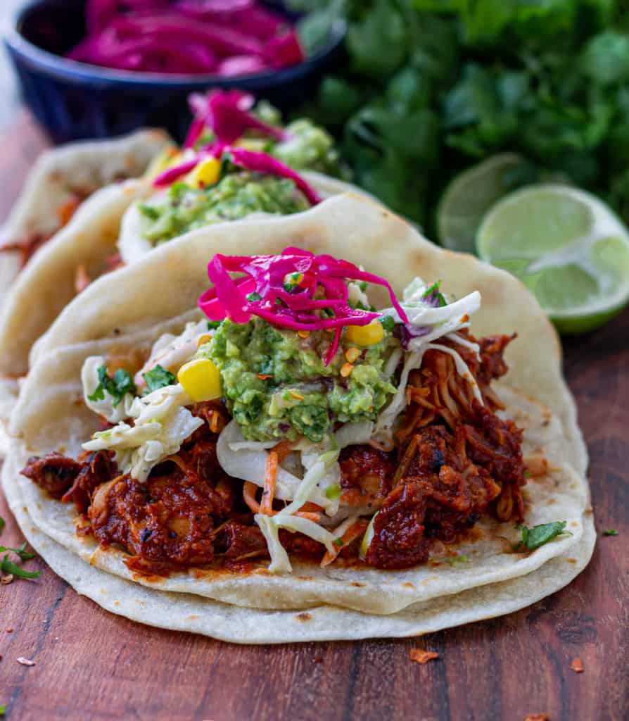 Three jackfruit tacos topped with coleslaw, guacamole, and pickled onions served on a tray.