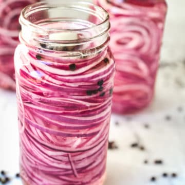 Three jars of pickled red onions on a board with black peppercorns scattered on counter.