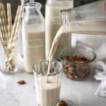 Fresh almond milk being poured into a glass on a tray with many jugs of milk and a bowl of almonds.