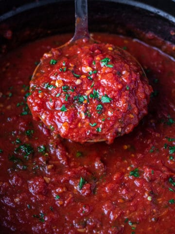 A close up ladleful of rich red marinara sauce being pulled out of a pot of sauce.