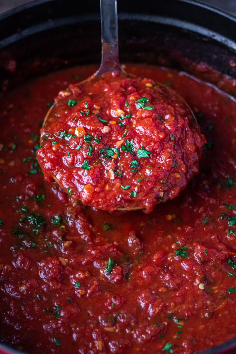 A close up ladleful of rich red marinara sauce being pulled out of a pot of sauce.