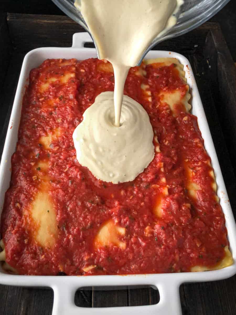 Vegan cheese sauce being poured on top of a pan of vegan lasagna.