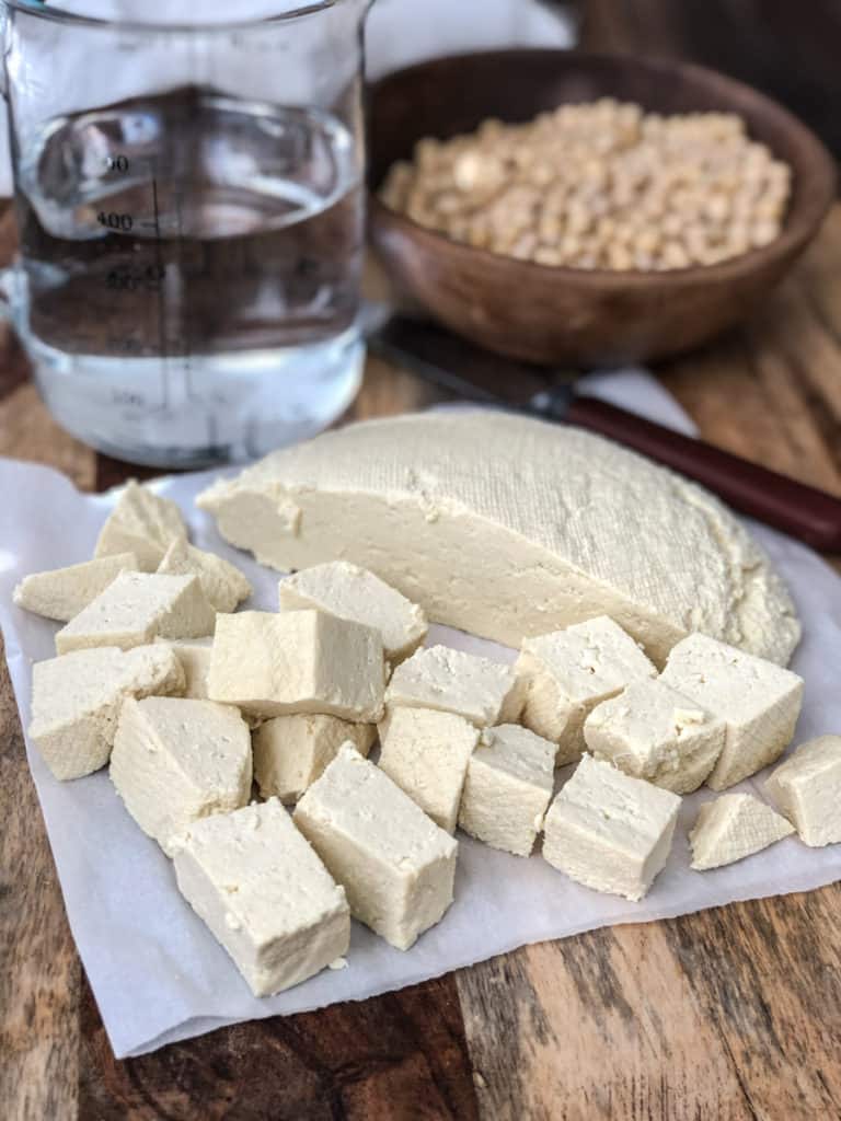 A block of freshly made tofu with some cut off on a paper towel in front of a bowl of soy beans and water.