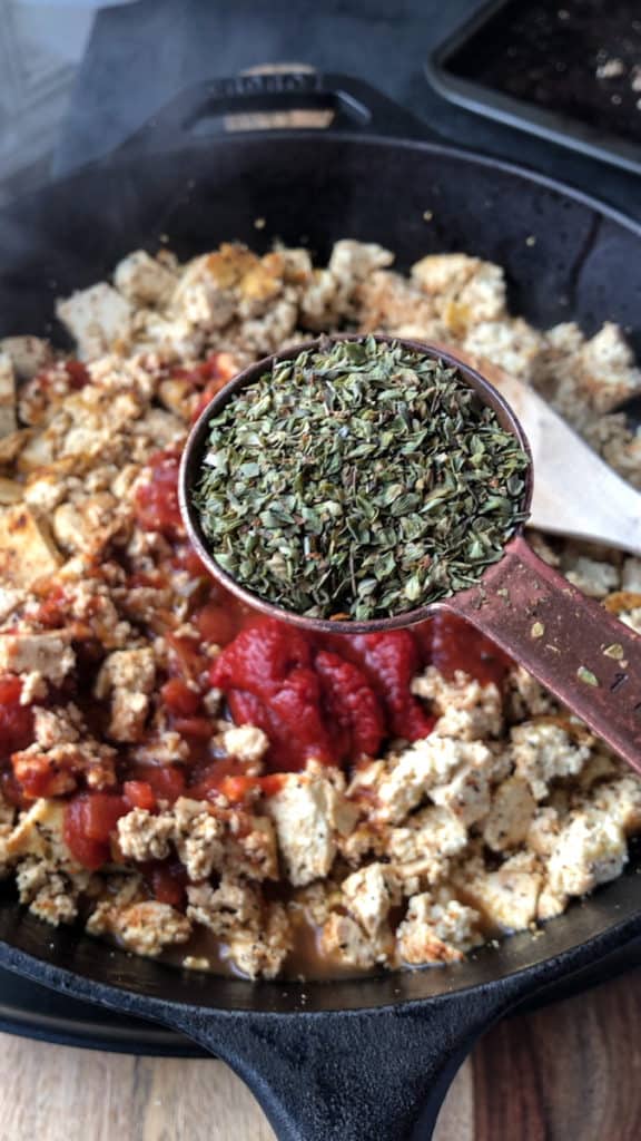 A tablespoonful of oregano being add to a frying pan of spicy tofu crumbles.