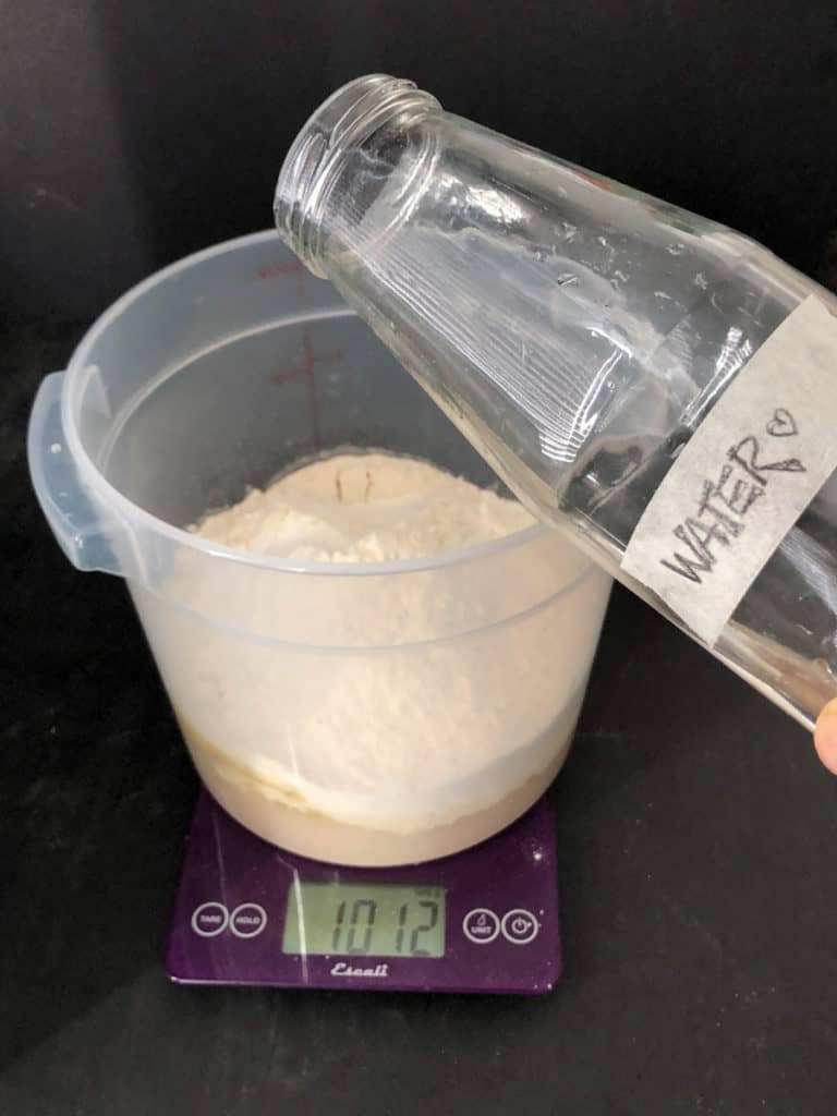 Bottle of water being poured into bucket of bread dough.