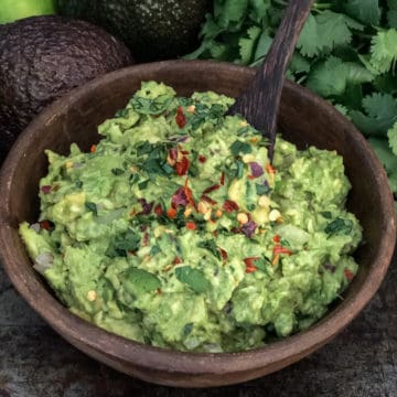 Bowlful of guacamole on a tray with avocados and cilantro.
