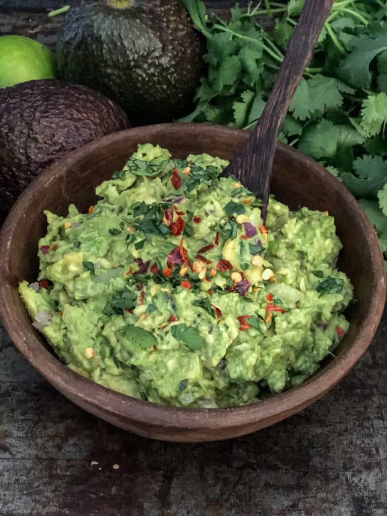 Bowlful of guacamole on a tray with avocados and cilantro.