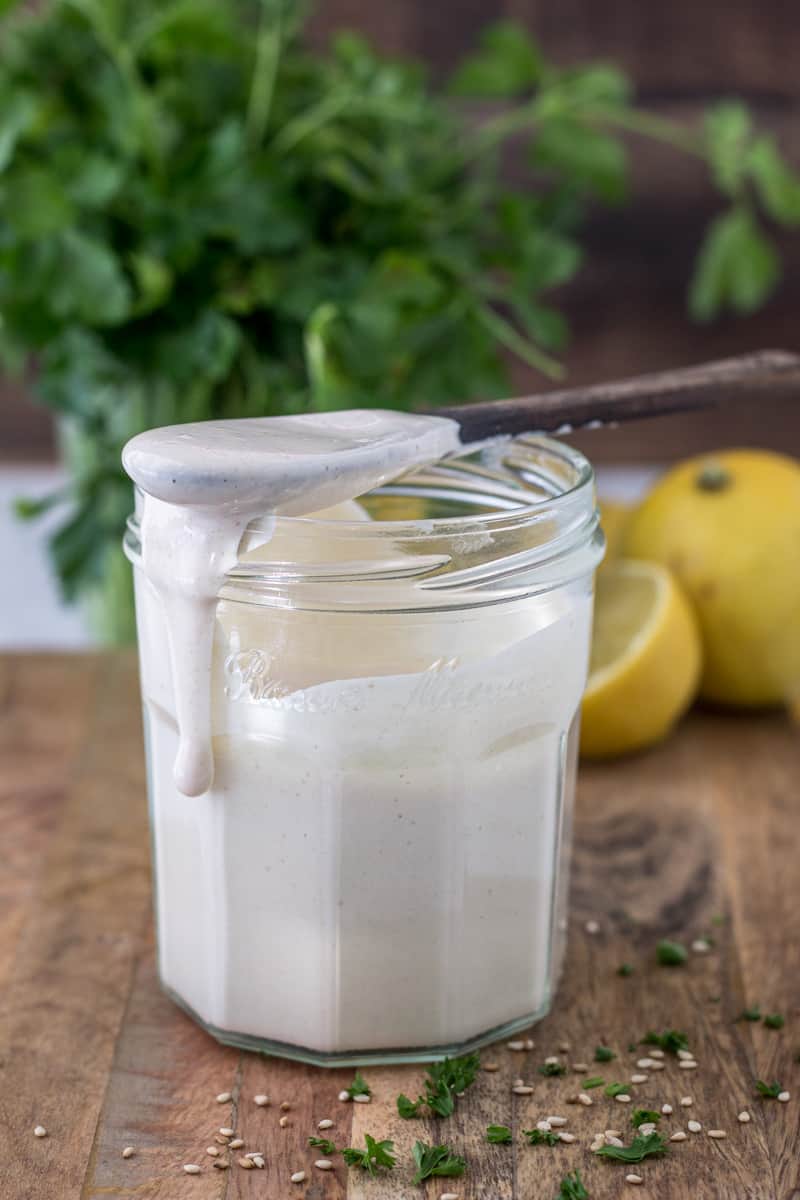 Jar of tahini sauce dripping out of jar with spoon resting on top.