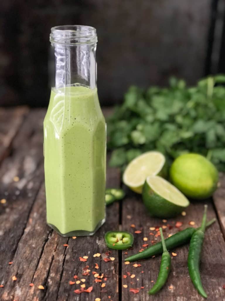 Jar of vegan green tahini sauce with limes and cilantro on a cutting board. 