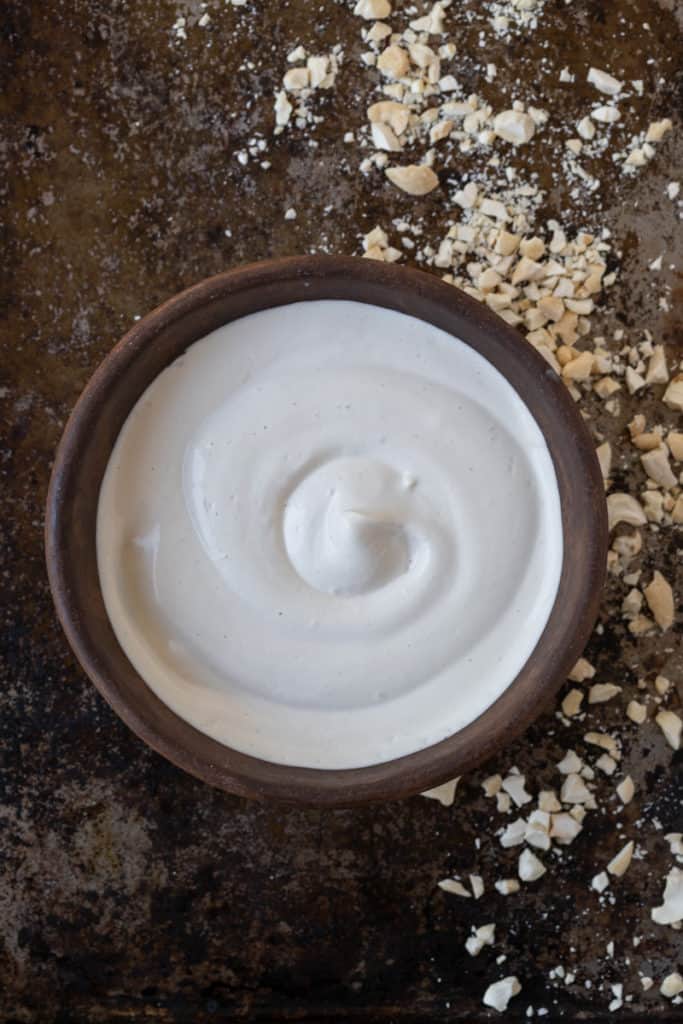 Bowl of vegan cashew cream sauce and crushed cashew nuts on a baking tray.