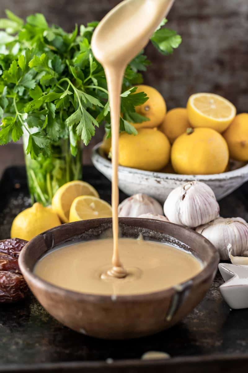 Tahini paste being drizzled into a bowl surrounded by lemons and garlic.