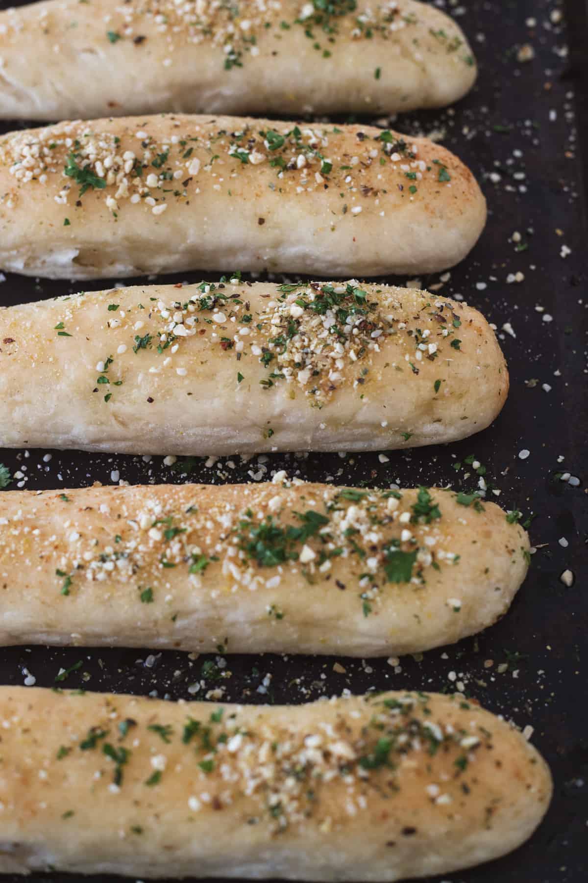 Baking sheet of golden breadsticks sprinkled with Italian herbs and parmesan.