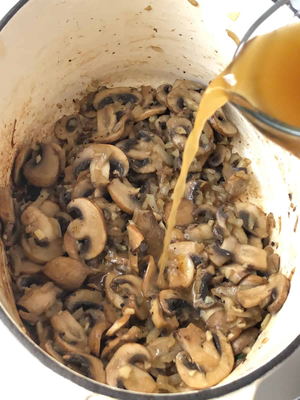 Vegetable broth being poured into a Dutch oven filled with sauteed mushrooms and onions.