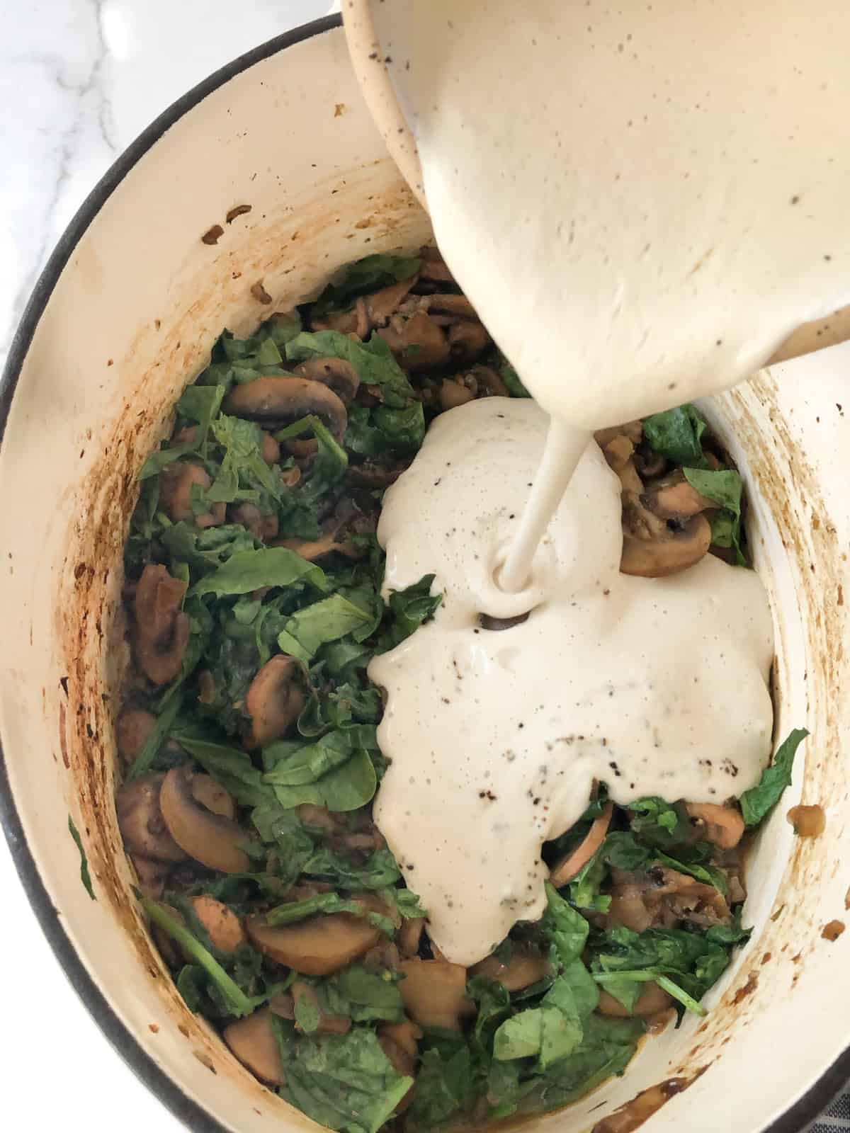 Cashew cream being poured into a Dutch oven filled with sauteed mushrooms and spinach.