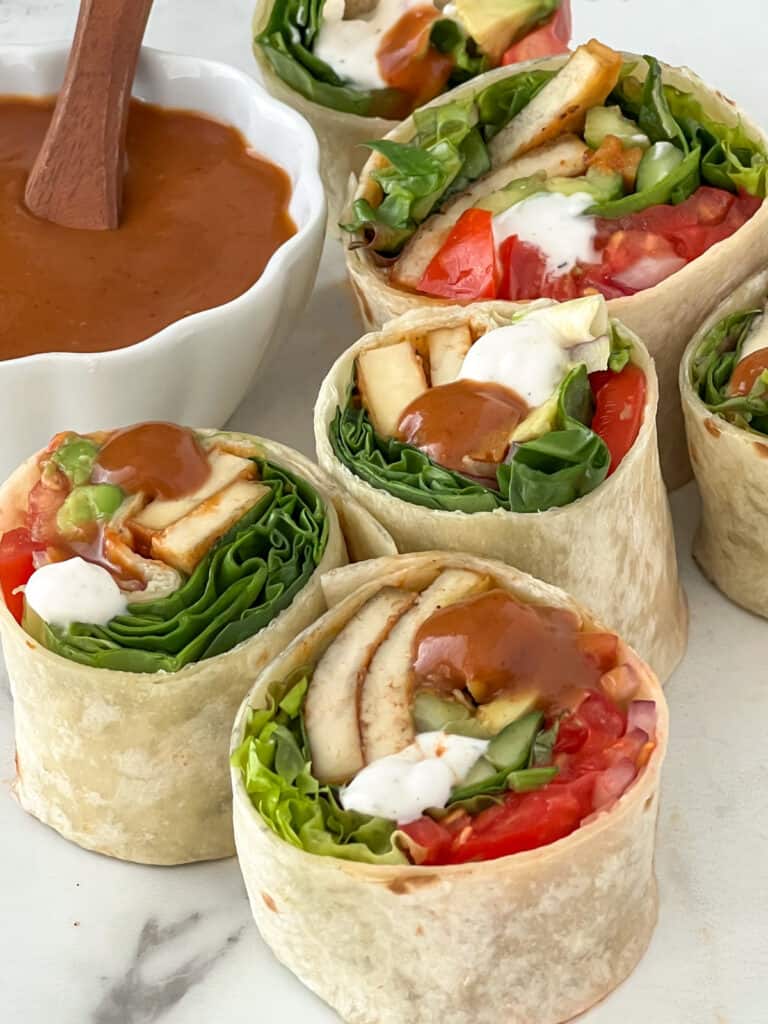Tofu and vegetable wraps cut in half on a cutting board with dish of BBQ sauce.
