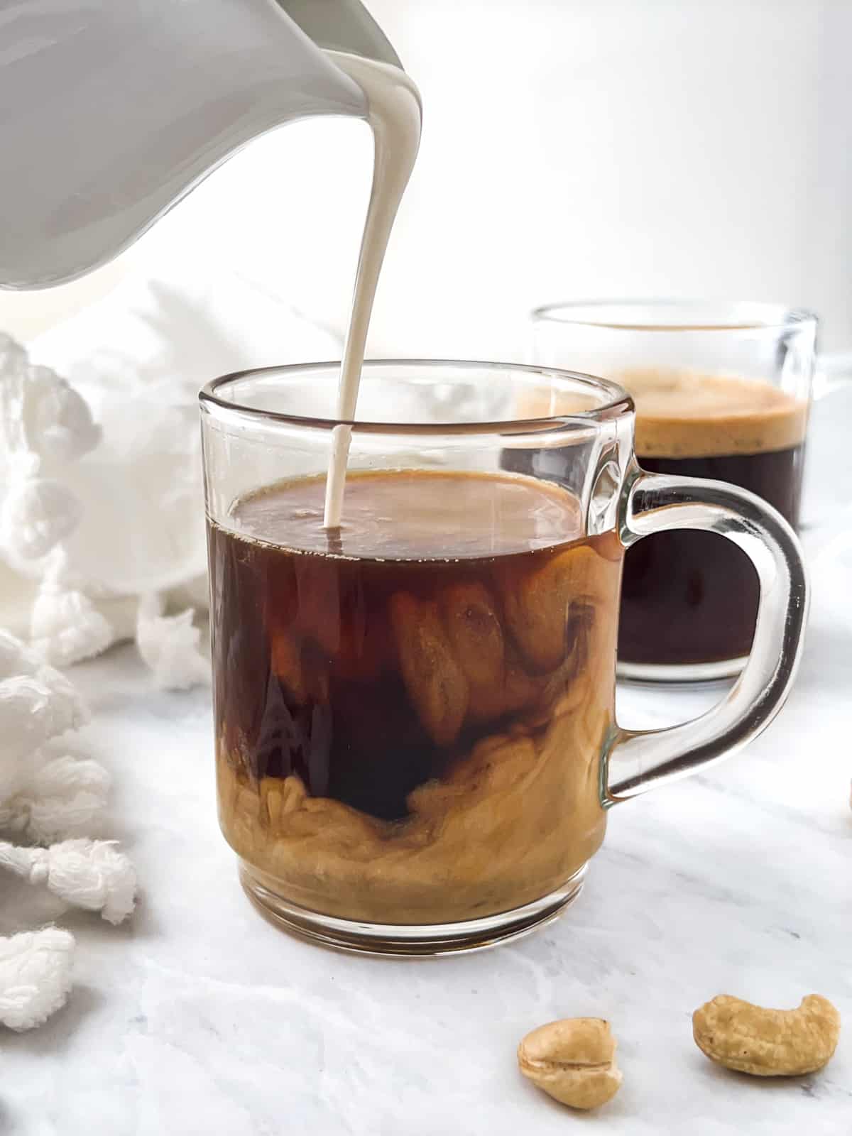 Heavy cream being poured into a coffee cup surrounded by white napkin and cashew nuts.