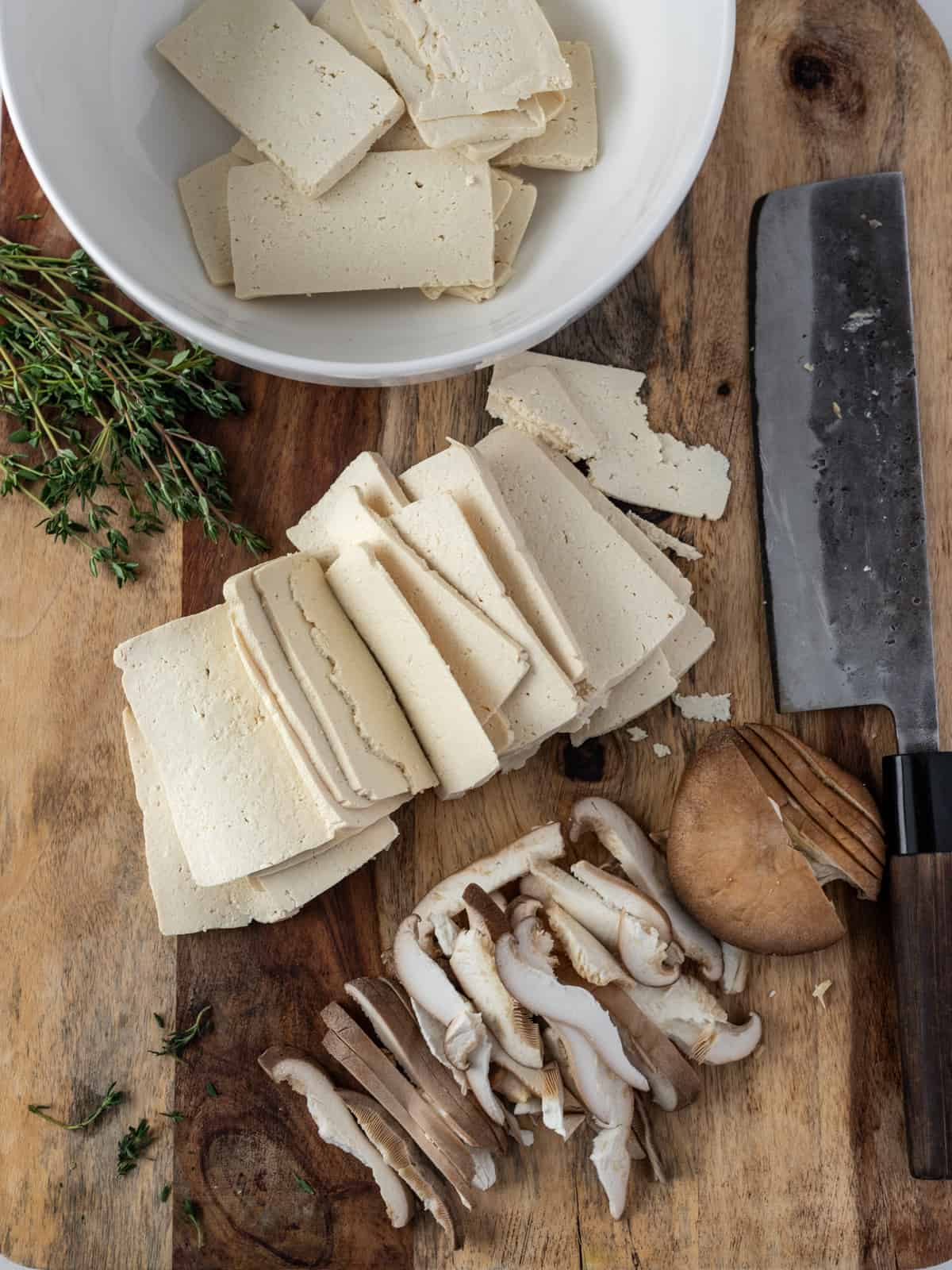 Slices of tofu in a white bowl and on a wooden cutting board with sliced mushrooms and a knife.