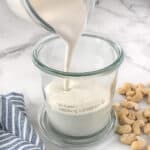 Vegan heavy cream being poured into a jar. Surrounded by cashew nuts and a blue and white striped napkin.