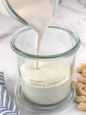 Vegan heavy cream being poured into a jar. Surrounded by cashew nuts and a blue and white striped napkin.