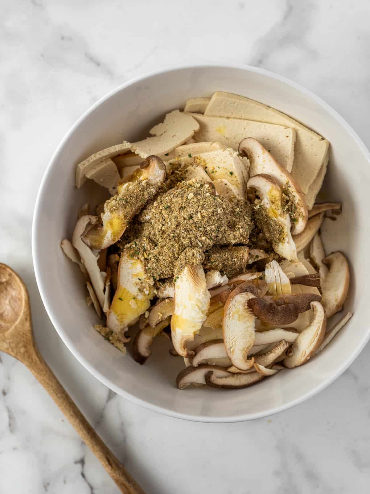 White bowl full of shaved tofu and sliced mushrooms on a white marble cutting board with a wooden spoon.