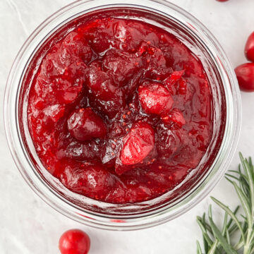 Bowlful of cranberry sauce surrounded by fresh cranberries and a sprig of rosemary.