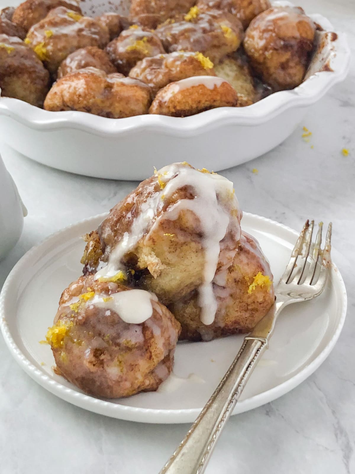 Cinnamon bites drizzled with icing sitting on a white plate in front of a pan of monkey bread.