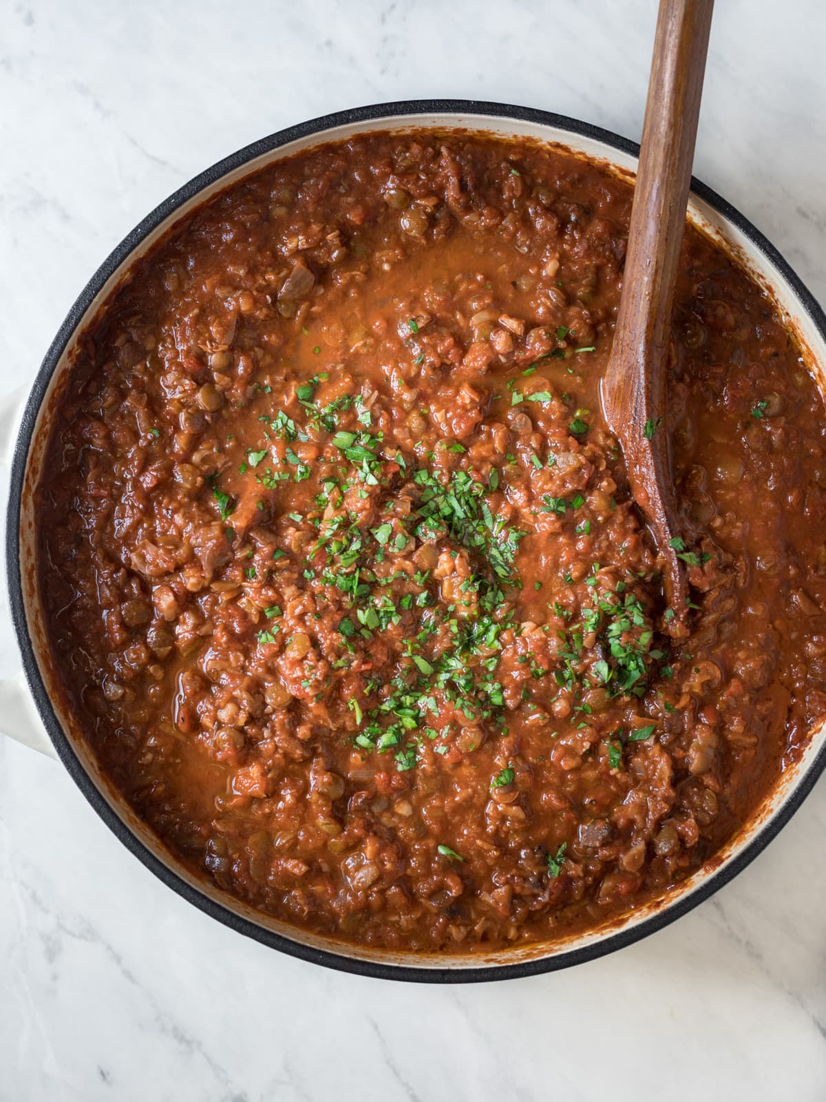 Large pot of lentil bolognese sauce topped with chopped parsley being stirred.