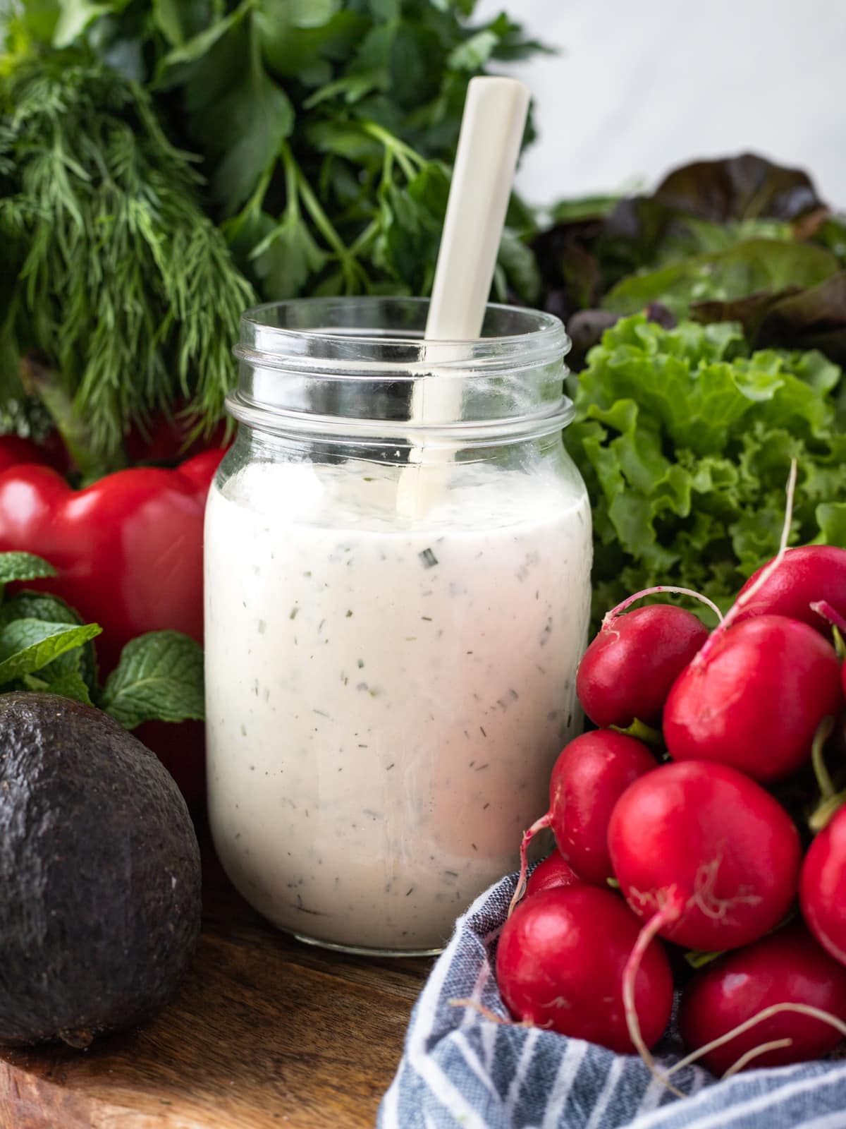 Jar of ranch dressing with a spoon surrounded by radishes, avocado tomatoes and lettuce greens.