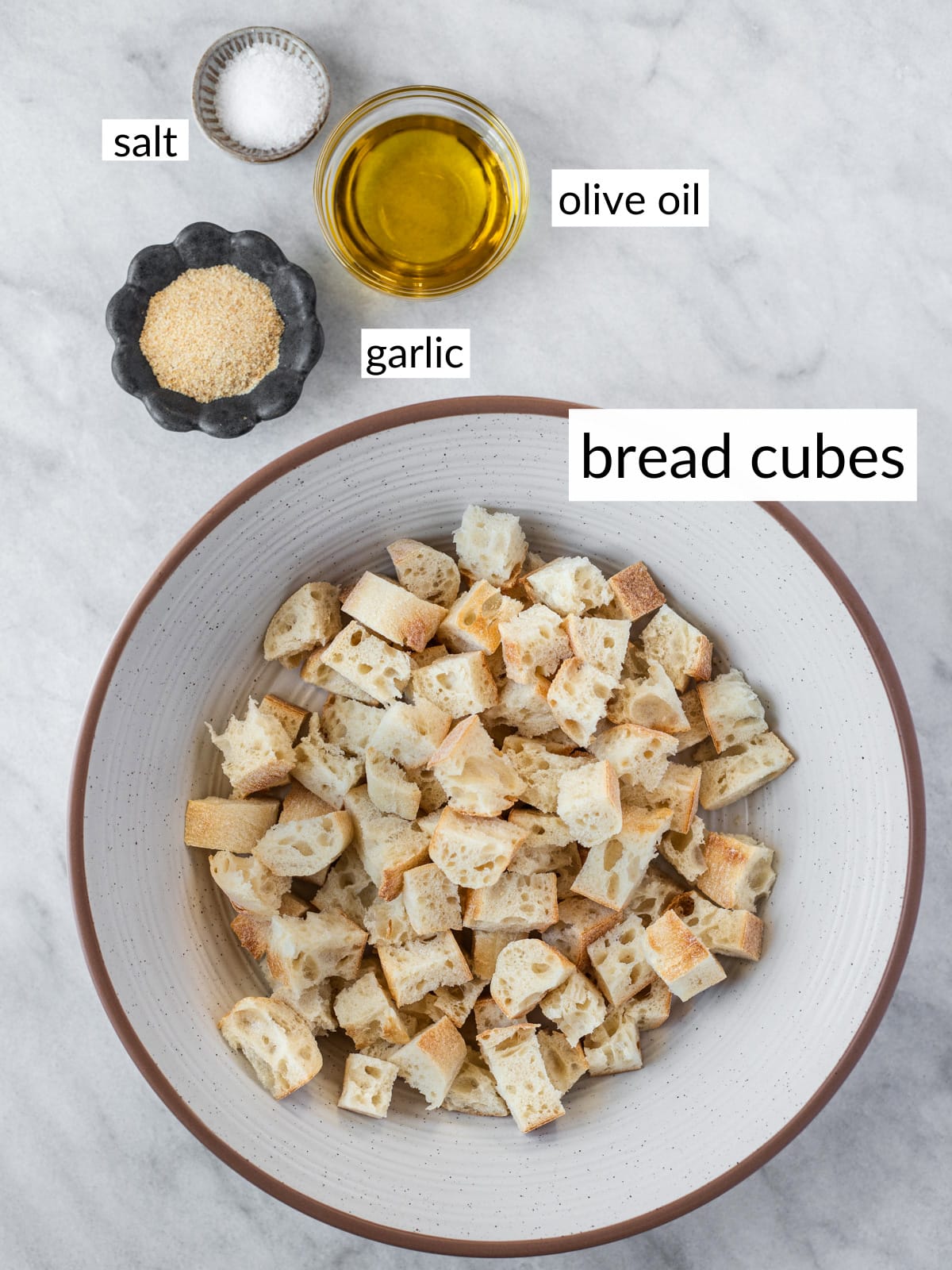 Ingredients to make croutons in bowls on a white marble counter.