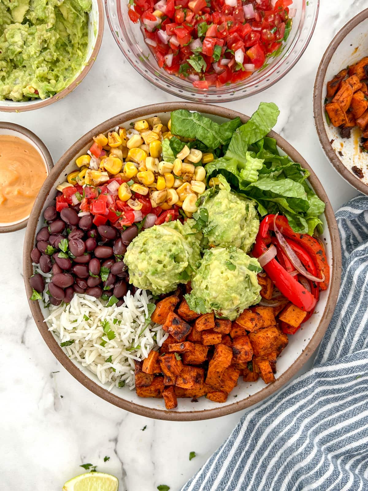 Vegetable burrito bowl surrounded by bowls of toppings including spicy cashew sauce, guacamole, pico de gallo, and fajita spiced onions and peppers.