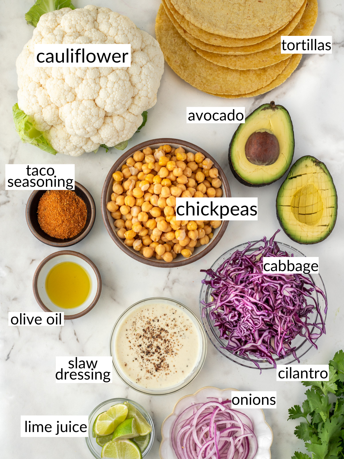 Ingredients for cauliflower chickpea tacos set out in bowls on the counter including avocado, cabbage and seasoning.