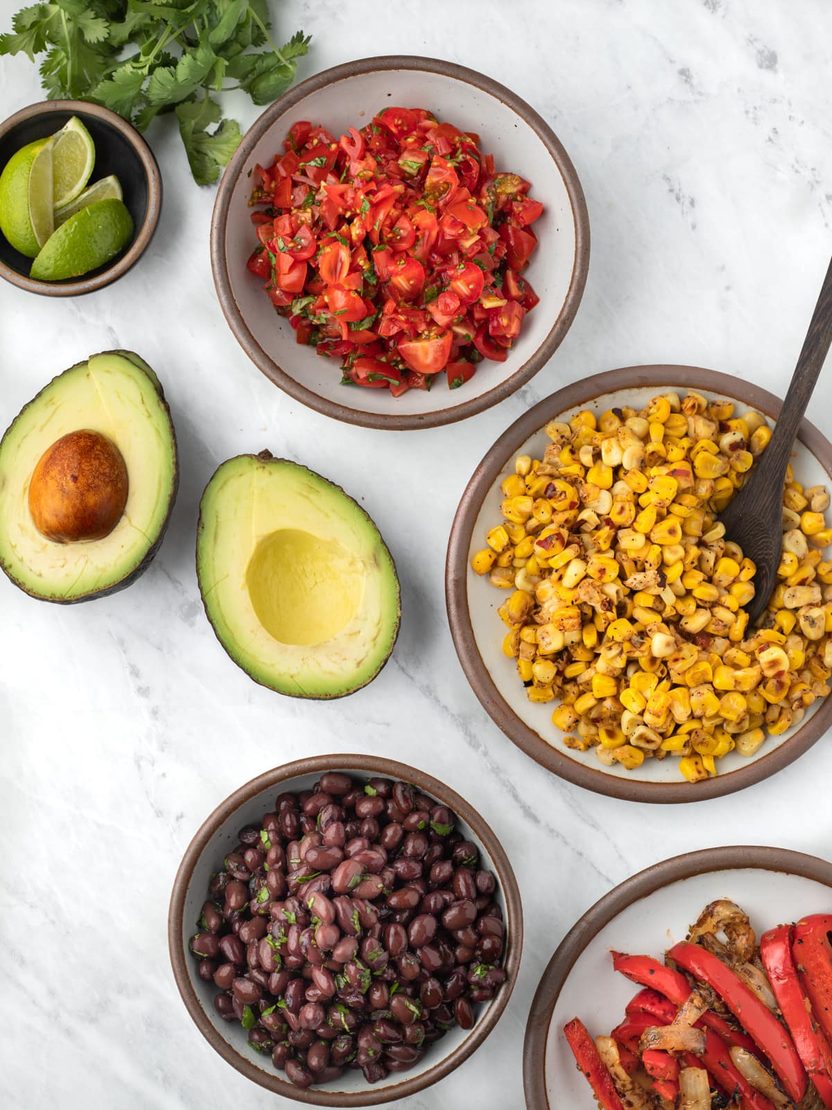 Bowls filled toppings for vegan burrito bowl: pico de gallo, spicy grilled corn, black beans, avocado halves,and fajita spiced peppers and onions.
