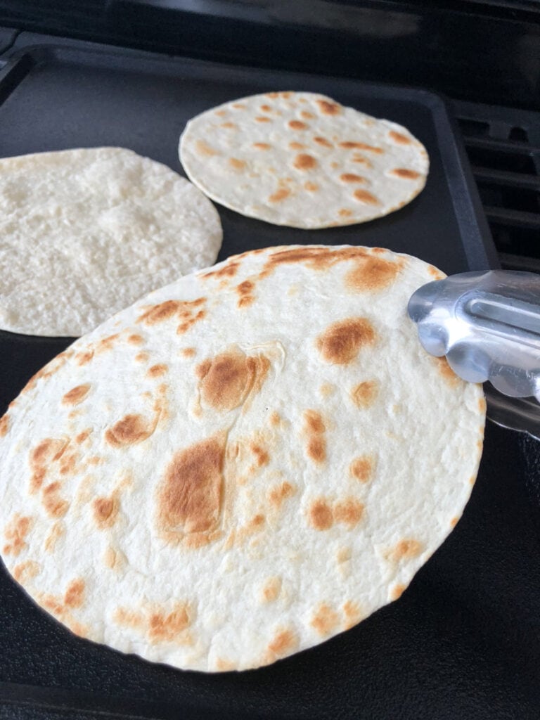 A warm tortilla being flipped with tongs on a griddle.