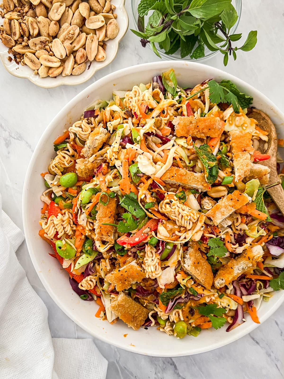 Overhead shot of Chinese chicken salad in a bowl surrounded by fresh herbs and roasted peanuts.