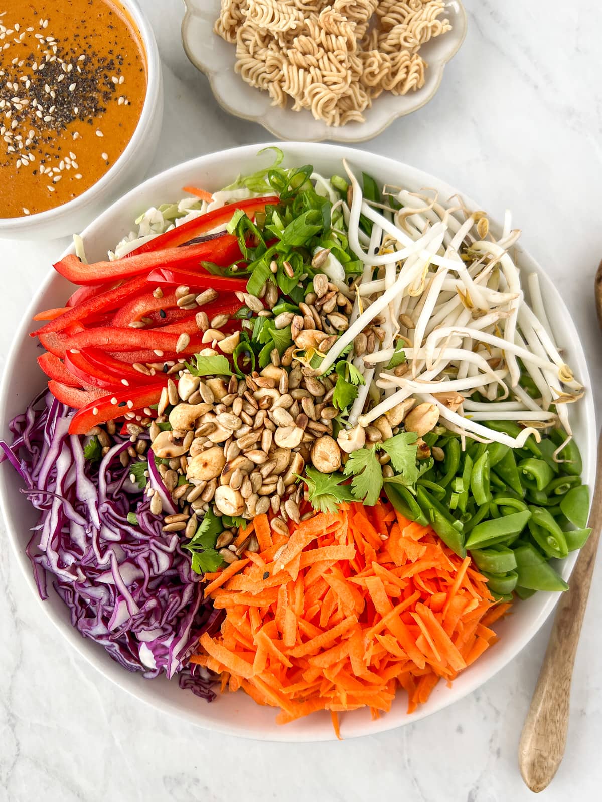 Ingredients to make an Asian chopped salad in a large serving bowl surrounding by smaller bowls of toasted ramen noodles and Asian carrot ginger dressing.