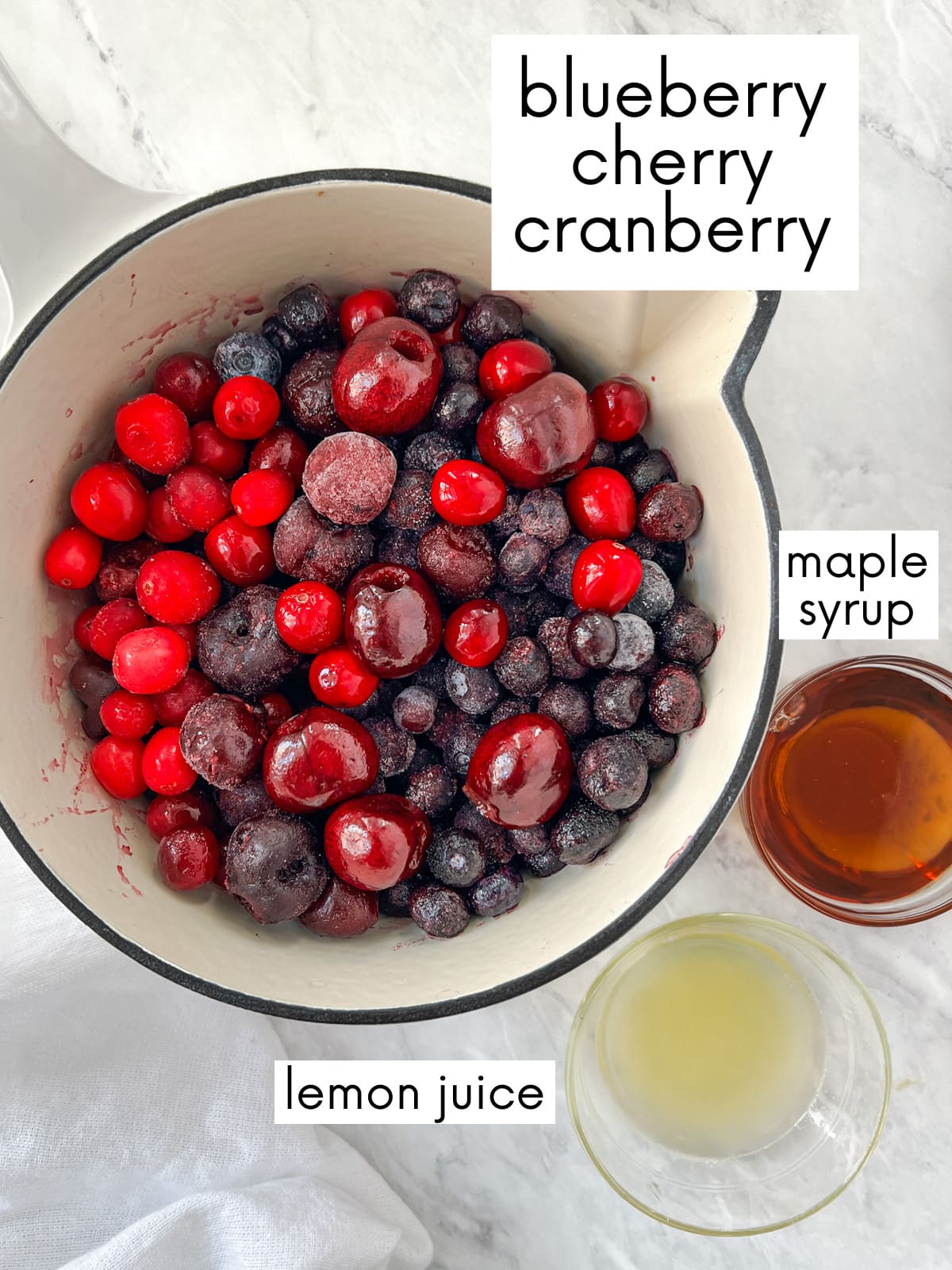 Sauce pan full of blueberries, cherries and cranberries along with small dish of lemon juice and maple syrup for making homemade jam.