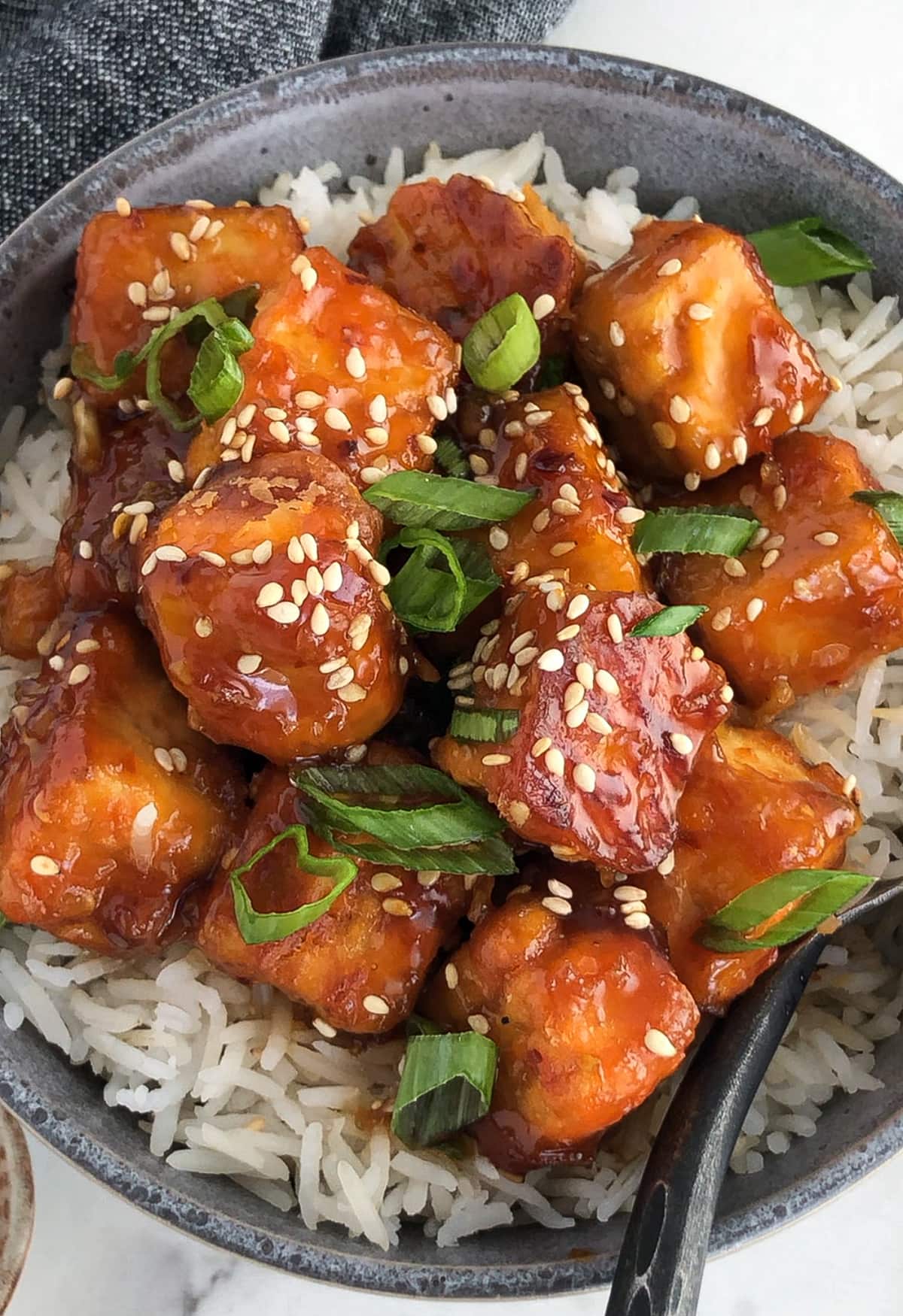 Crispy sesame garlic tofu served on rice and garnished with sliced green onions and sesame seeds.