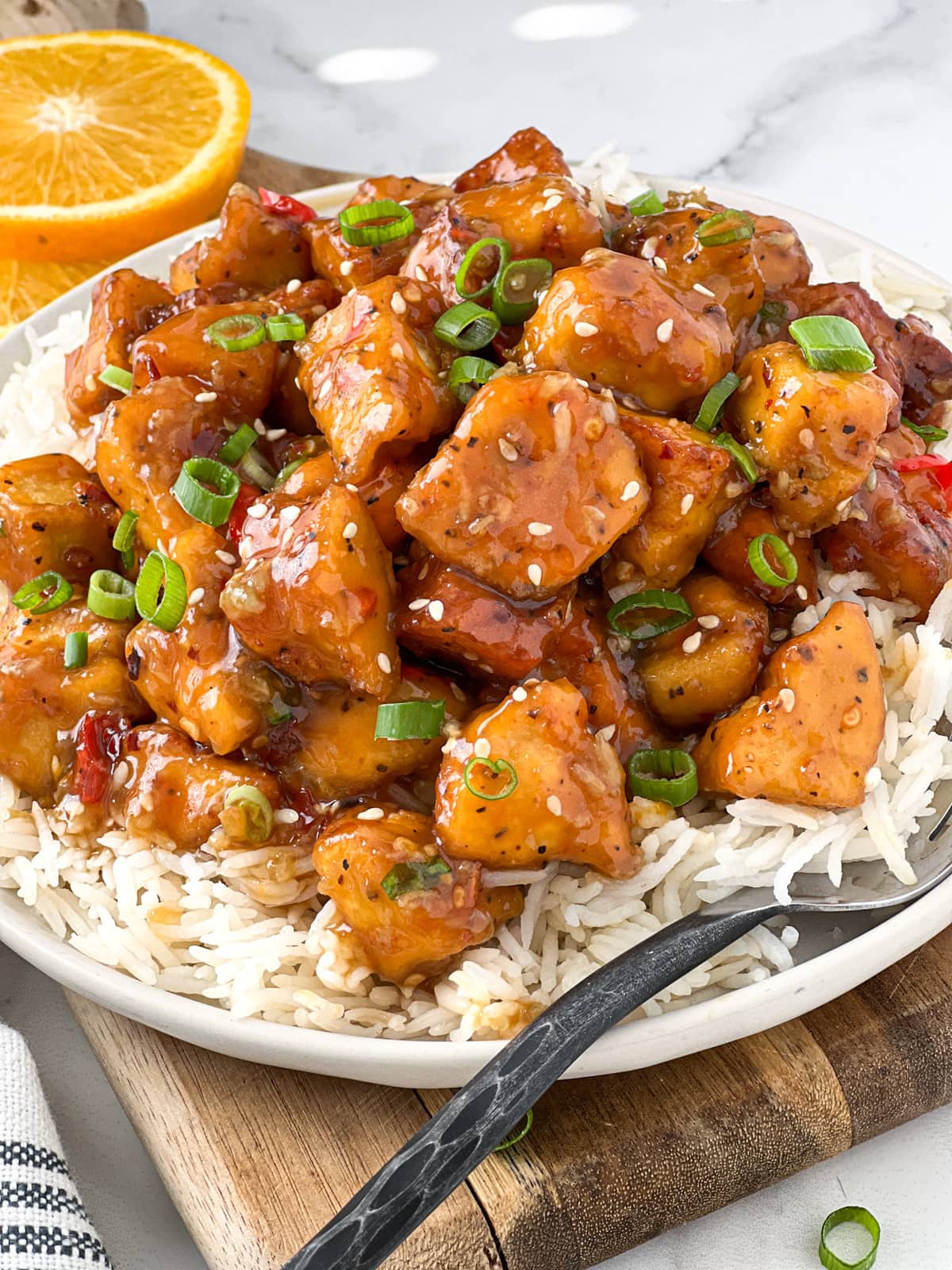 Plate of saucy orange tofu served on a bed of rice with a sprinkle of green onions and sesame seeds.