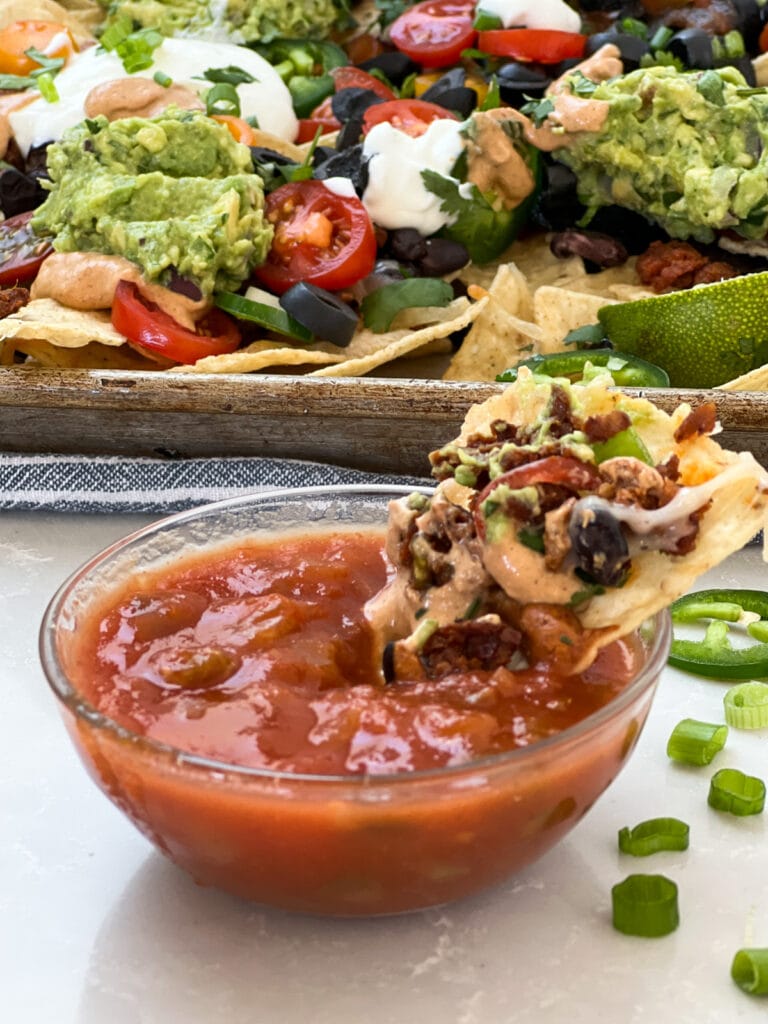 Loaded vegan nacho being dipped into bowl of salsa.